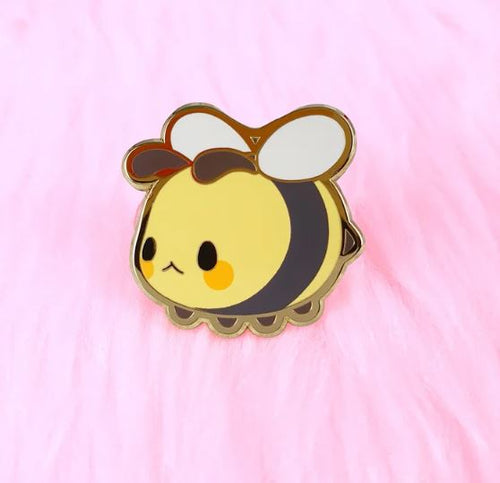 A charming and plump Bee Pin Pinstachiio by Blue Dog Posters, featuring large white wings, a cute face with round black eyes and yellow cheeks. This hard enamel pin rests on a soft, fluffy pink surface for an added touch of appeal.