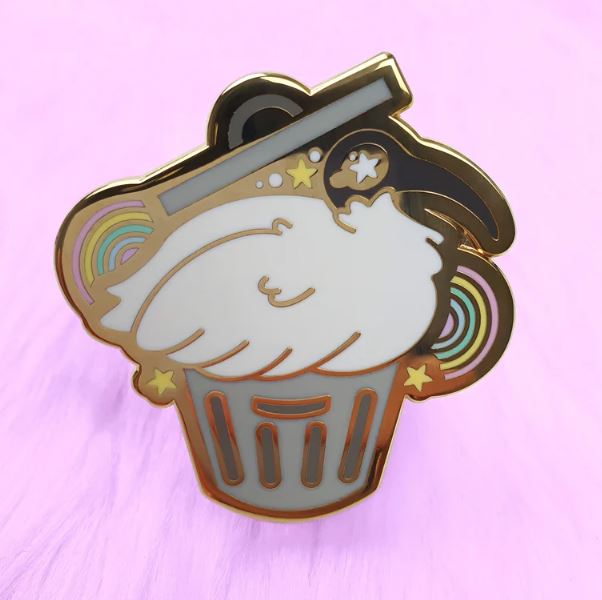The Bin Chicken Pin Pinstachiio by Blue Dog Posters is a gold-plated enamel pin that features a white frosted cupcake with gold edges on a light purple background. Vibrant rainbow accents and stars surrounding the cupcake add a whimsical touch. It includes rubber clutches for secure fastening.