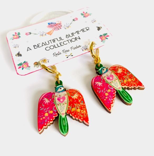 Colourful bird-shaped earrings from Blue Dog Posters' "A Beautiful Summer Collection" hang on a card. These vibrant earrings feature shades of pink, green, and orange with intricate patterns.