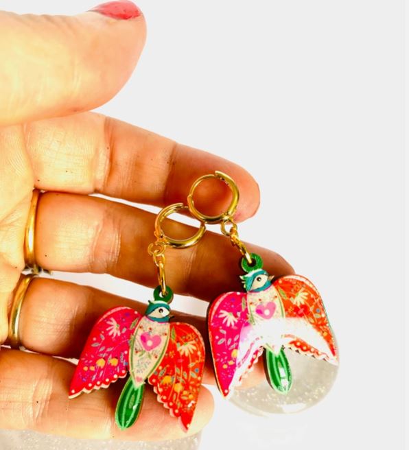 A hand displays Blue Dog Posters' Colourful Bird Dangle Earrings, showcasing vibrant red, pink, and green hues with intricate patterns. The earrings feature gold hoop closures against a plain white background.