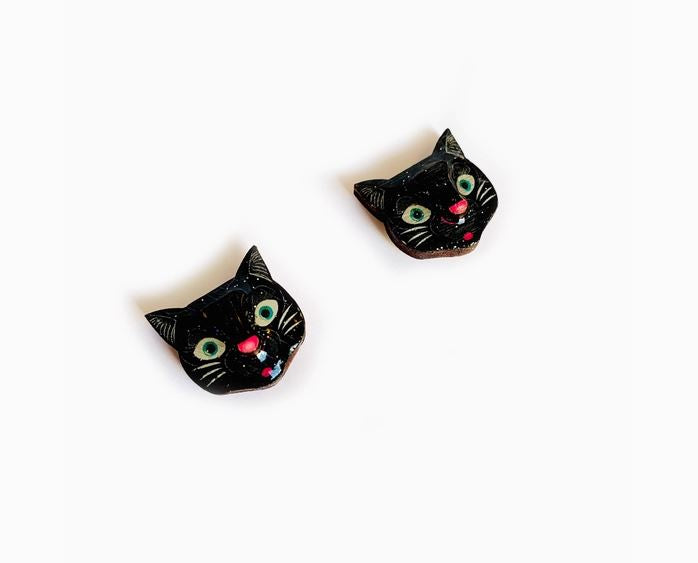 The Black Cat Stud Earrings by Blue Dog Posters feature a pair of black cat-shaped earrings with green eyes and pink noses, set against a white background. These earrings include small colorful details that give them character.