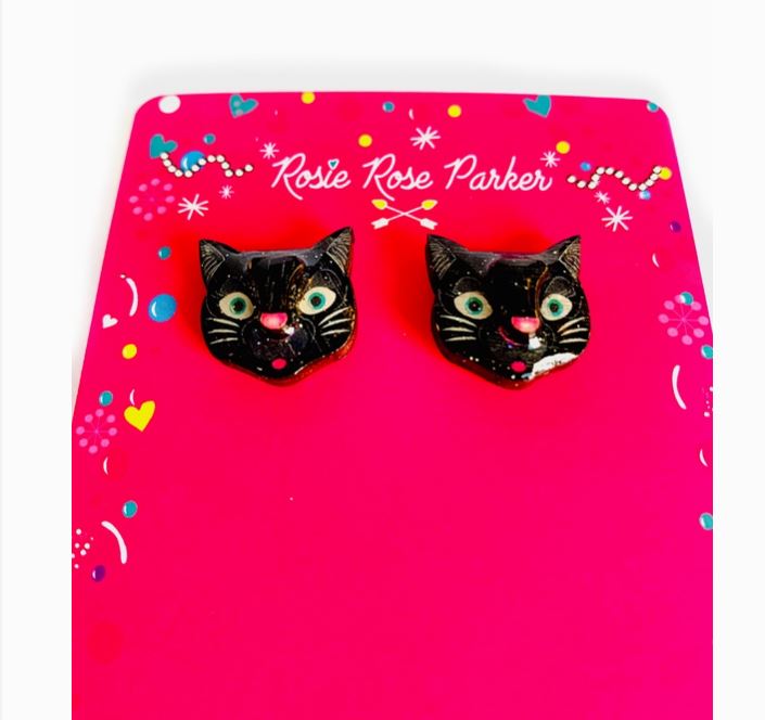 Black Cat Stud Earrings, designed as black cat heads with green eyes and pink noses, are showcased on a bright pink cardboard backing featuring "Blue Dog Posters" and decorative hearts and swirl patterns.