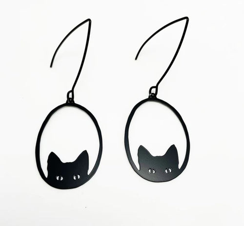 Introducing the Denz Mini Earrings Black Cat by Blue Dog Posters: Each pair of these lightweight earrings is crafted from recycled plated stainless steel, showcasing a minimalist and whimsical design. These earrings feature a simple, black silhouette of a cat head with pointed ears and circular cut-out eyes, all enclosed within a thin, black oval frame.