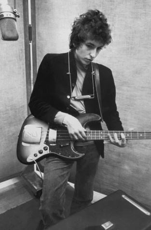 In the Bob Dylan Studio by MUS 284, a man with curly hair plays electric bass near a microphone, fully focused on his instrument. A large wall poster adds creative flair to the recording space.