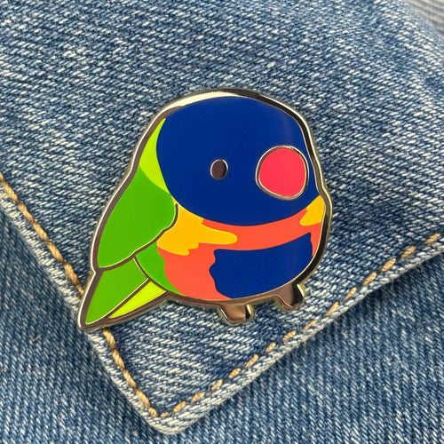 A Lorikeet Pin Pinstachiio by Blue Dog Posters, showcasing a vibrant palette of blue, green, red, and orange in a cute, cartoonish design with a gold-plated finish, is attached to a denim jacket. For collectors' assurance, it includes an authenticity back-stamp.
