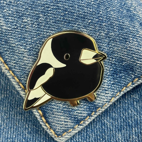 A black and white enamel pin named "Magpie Pin Pinstachiio" from Blue Dog Posters, featuring a small, cute bird with minimalistic details, is securely attached to a piece of blue denim fabric with visible stitches. For an added touch of elegance, the pin is gold plated.