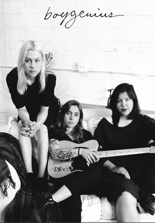 A black and white photo featuring three women from the band Boygenius, sitting on a couch. One member stands and leans forward slightly, while the other two sit; one holds a guitar. The band's name is handwritten above them on the brick wall. Perfect for any music collection or as an indie rock poster. Product Name: Boygenius by MUS 150.