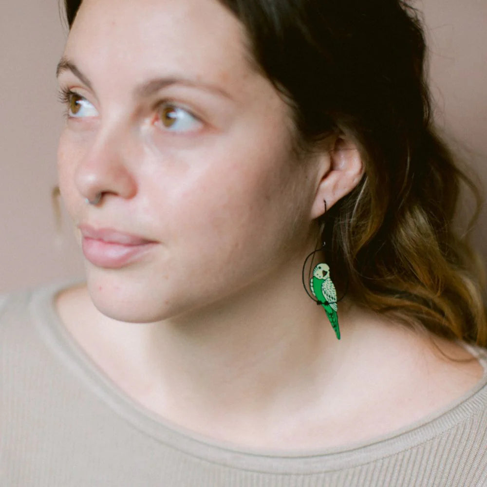 A woman with wavy brown hair wears a light top and Blue Dog Posters' Budgie Black Green Yellow Denz Earrings, shaped like green parrots. She gazes softly to the left against a neutral background.