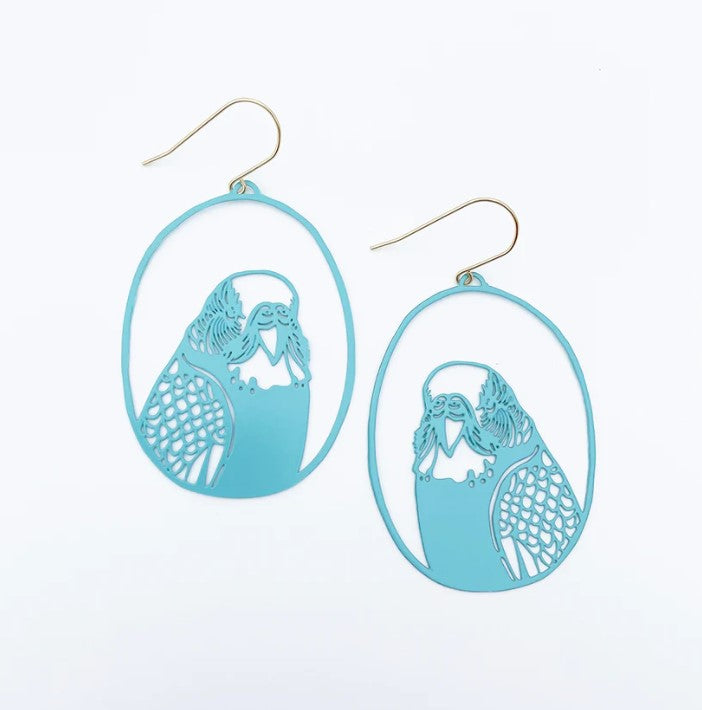 The Budgie Dangle Earrings by Blue Dog Posters are a pair of light blue earrings featuring intricate cutout designs of budgies, each set within an oval frame. These painted steel dangles have gold-colored hooks and are displayed against a plain white background.