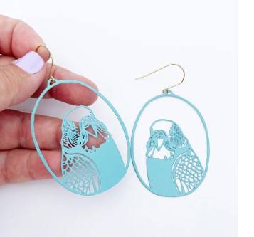 A hand holds a pair of Budgie Dangle Earrings by Blue Dog Posters. These light blue, oval-shaped earrings feature cutout designs of two parrots facing each other, and they are suspended by gold hooks. Made from recycled stainless steel, the earrings are photographed against a white background with light purple painted fingernails.