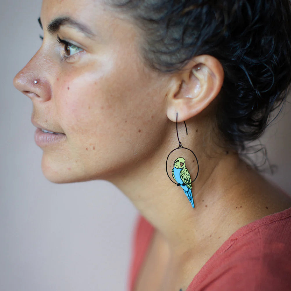 A person with short, curly dark hair and a nose piercing is seen in profile, wearing Blue Dog Posters’ Budgie Black Blue Yellow Denz Earrings shaped like a colorful parakeet and a coral-colored top.