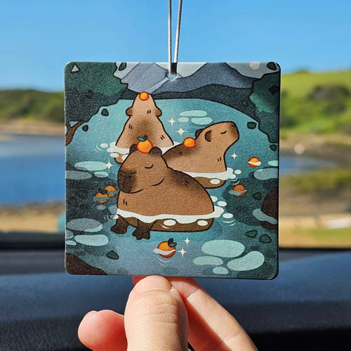 A hand holds the Capybara Orange Air Freshener - Pinstachiio by Blue Dog Posters, featuring cartoon Muscle Capybaras relaxing in a hot spring with oranges on their heads, hinting at the refreshing orange scent.
