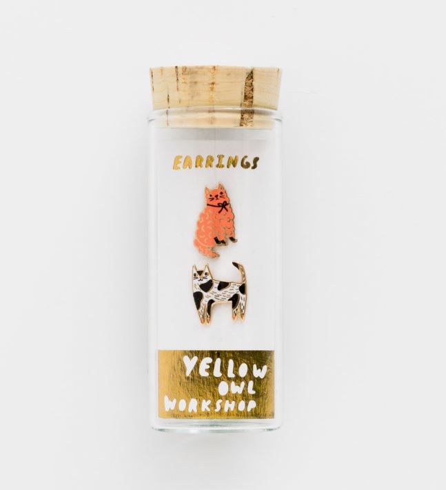 The image depicts a glass vial container with a cork lid, housing a pair of mismatched earrings. One earring features the shape of an orange poodle, while the other is designed as a black and white cat. Text on the vial reads "Cats Earrings" and "Blue Dog Posters" on a gold background, emphasizing hypoallergenic posts.
