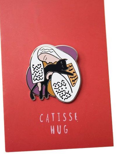 The Catisse Hug Enamel Pin by Blue Dog Posters is displayed on a red card. The high-quality pin, inspired by a Henri Matisse caricature, features a person wrapped in a blanket, hugging a black cat. The background includes abstract shapes in purple. Below the pin, the card reads "CATISSÉ HUG" in white letters.