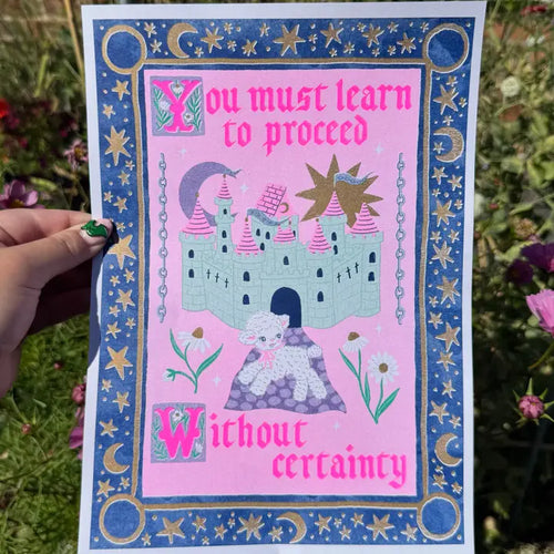 A hand holds the "Proceed Without Certainty A4 Risograph" by Amy Hastings from Blue Dog Posters, a vibrant medieval-themed print with castle, sun, moon, and sheep. Text: “You must learn to proceed without certainty.” Printed on recycled coffee cup paper.