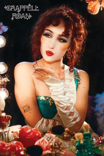 A woman with curly red hair and striking makeup blows a kiss. Clad in a teal and white ensemble featuring lace-up sleeves, she's surrounded by cosmetics and lights. The words "MUS 351 - Chappell Roan" appear in the corner.