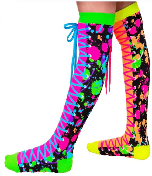 Two legs adorned with knee-high Madmia Colour Run Socks from Blue Dog Posters, showcasing a dynamic lace-up design and vibrant hues. The socks boast a black backdrop splashed with lively colors including pink, green, orange, blue, and yellow. Under UV light, the neon shoelaces—one in blue and one in pink—shine brightly. The toes are highlighted in green and yellow.