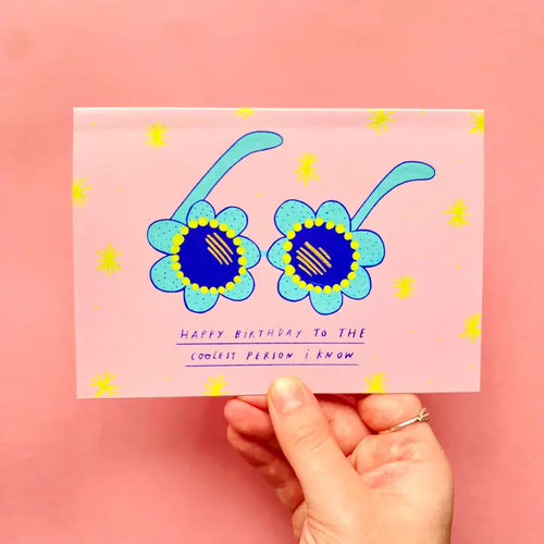 A hand holds Blue Dog Posters' Coolest Person I Know Greeting Card, featuring two blue and yellow flowers and the text "Happy birthday to THE coolest person I know," against a pink background with yellow starbursts.