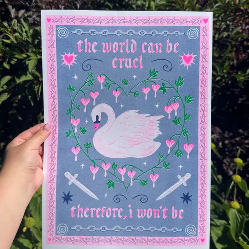 A hand holds "The World Can Be Cruel A3 Risograph" print by Amy Hastings from Blue Dog Posters, showing a white swan, pink hearts, and two swords with the text “the world can be cruel therefore, i won’t be.” Green foliage is in the background.
