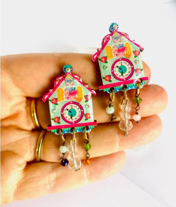 A hand holds two Cuckoo Clock Retro Earrings by Blue Dog Posters, each adorned like miniature cuckoo clocks with vibrant decorations and beads in assorted colors and shapes.