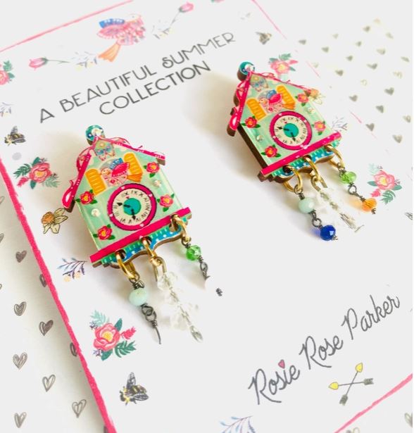 Colorful Cuckoo Clock Retro earrings feature house designs with clocks, vibrant beads, and charms. Displayed on decorative paper, they are part of the "A Beautiful Summer Collection" by Blue Dog Posters in collaboration with Rosie Rose Parker.