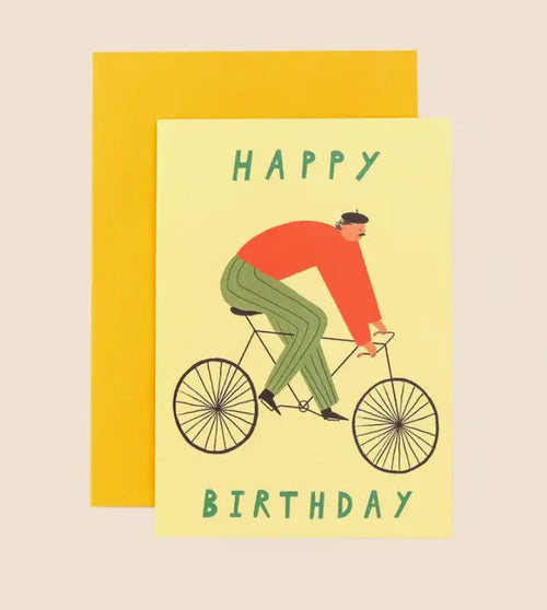 The Cyclist Happy Birthday Greeting Card from Blue Dog Posters showcases a delightful illustration by Little Black Cat Illustrated, depicting a person wearing a red shirt, green pants, and a black hat while riding a bicycle. The words "HAPPY BIRTHDAY" are elegantly placed above and below the design. Set against a beige background and paired with an orange envelope, this charming card is crafted in Manchester, UK.