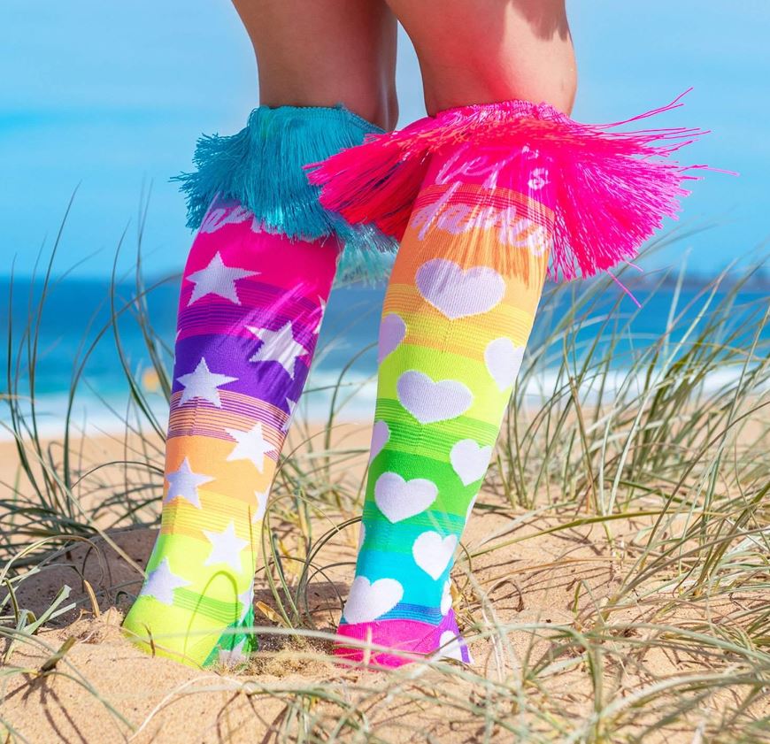 A person stands on sandy dunes by the beach, wearing vibrant Madmia Lets Dance Socks from Blue Dog Posters. These knee-high socks feature playful star and heart patterns in white set against a rainbow gradient background, topped off with unique pink and blue custom-made tassels. Grass and ocean waves can be seen nearby.