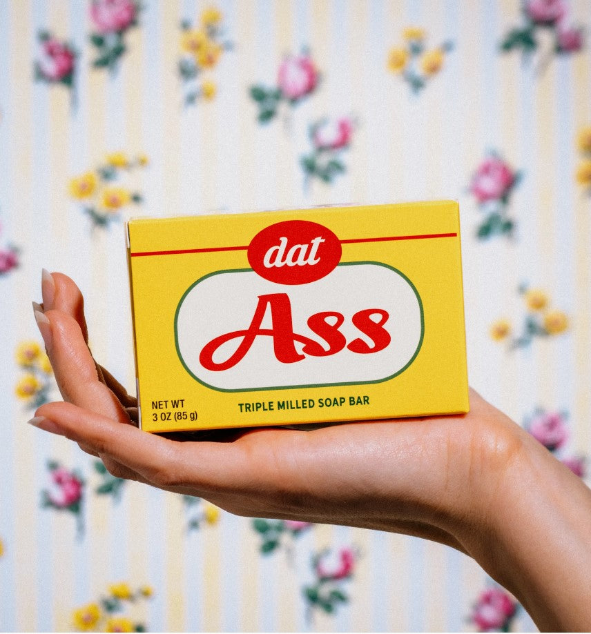 A hand holds a yellow soap bar package featuring "dat" in small red text above "Ass" in large red text. The decorative background includes vertical yellow stripes and pink roses. This 3 oz (85 g) Vermont-made soap, named Dat Ass Triple Milled Boxed Bar Soap, is from Blue Dog Posters.