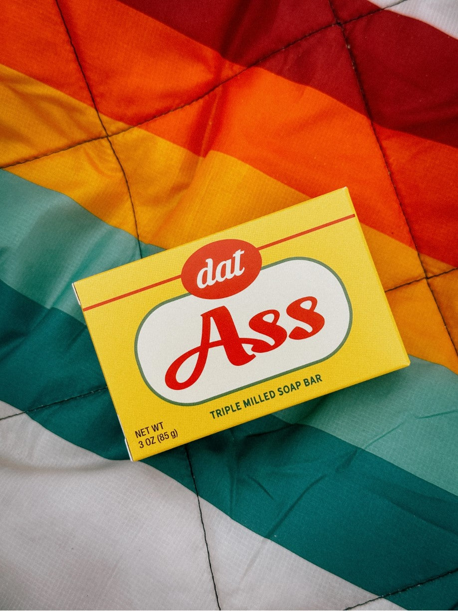 A yellow soap bar box from Blue Dog Posters labeled "Dat Ass Triple Milled Boxed Bar Soap," weighing 3 oz (85 g). The Vermont-made soap box is placed on a colorful quilt with diagonal stripes in red, yellow, orange, green, and teal.