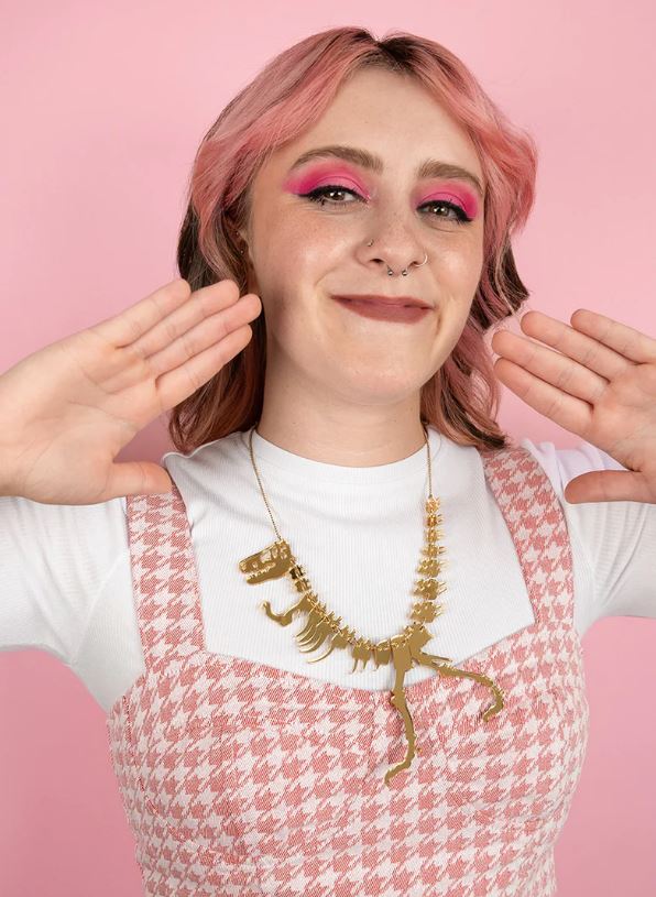 A person with wavy pink hair and bright pink eyeshadow smiles against a pink background. They wear a white shirt paired with a Blue Dog Posters Dinosaur Necklace in Gold Colour and a pink houndstooth dress, hands playfully raised as if ready for a Jurassic adventure.