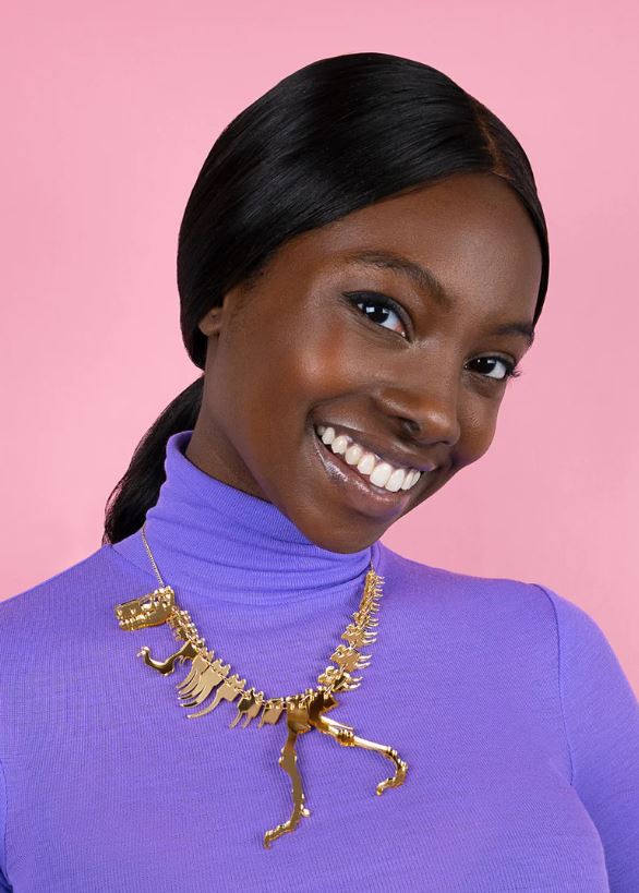 A person with long dark hair, smiling, is wearing a purple turtleneck and a striking gold dinosaur-themed necklace from Blue Dog Posters. The background is pink.