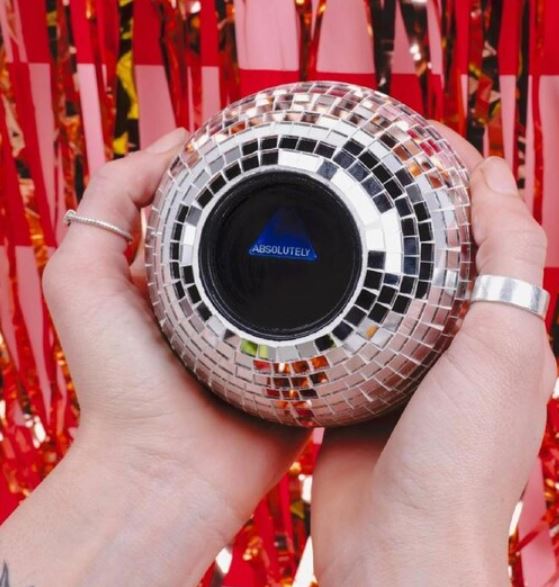 A person holds the Fizz Creations Disco Decision Ball by Blue Dog Posters, showing "ABSOLUTELY" with red and pink shiny streamers in the background—ideal as a quirky decision-making toy or fun party décor.
