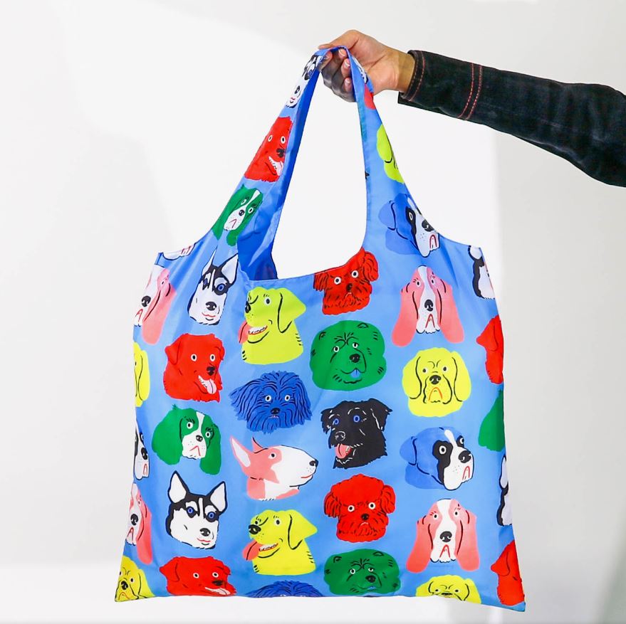 A person is holding a Dogs Art Sack by Blue Dog Posters, showcasing a colorful reusable bag adorned with illustrations of various dogs on a light blue background. Designed by Kristina Micotti and crafted from 100% recycled rPet, the bag features dogs of different colors and breeds, creating an energetic and playful design. The person's hand is visible from the wrist down, dressed in a dark sleeve.