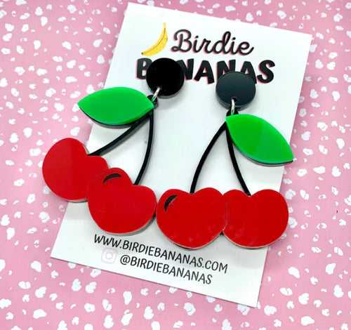 A pair of Double Cherry Acrylic Earrings featuring vivid red cherries and green leaves is showcased on a white card adorned with "Blue Dog Posters" branding. Handmade in Australia, this card sits against a pink background decorated with small white polka dots. The website and Instagram handle are clearly visible on the card, making it ideal for vintage fashion enthusiasts.