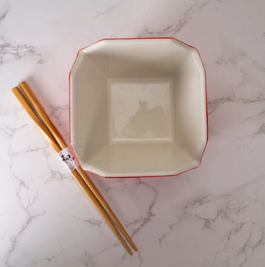 The Blue Dog Posters Takeout Box Serving Bowl With Chopsticks GOLD DRAGON, a stylish and microwave-safe white ceramic bowl with a thin red rim, sits on marble beside wooden chopsticks in a paper sleeve—perfect for everyday dining.