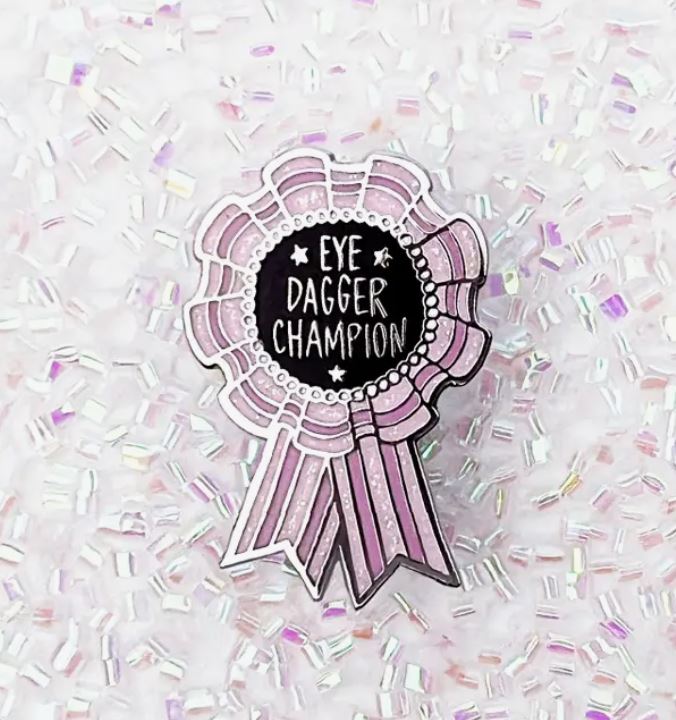 A pink and white ribbon-shaped hard enamel pin with the words "Eve Dagger Champion" written in white on a black circular center. This cool pin, created by Blue Dog Posters, is surrounded by iridescent beaded decorations on a white background.