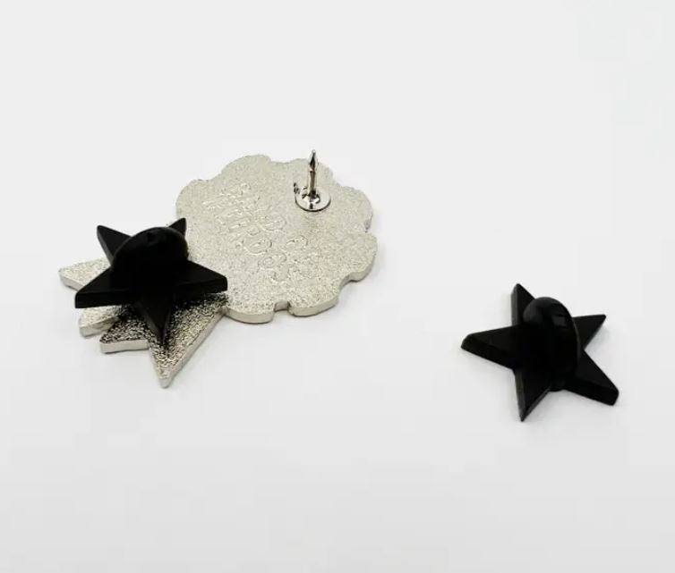 An Eve Dagger Champion Enamel Pin by Blue Dog Posters, featuring a black star shape crafted with hard enamel and a silver metal backing, lies face up, while another sleek pin rests face down on a plain white background.