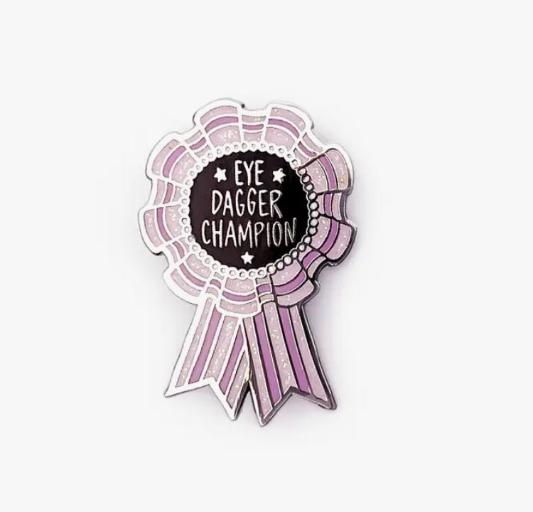 Introducing the Eve Dagger Champion Enamel Pin from Blue Dog Posters! Featuring a vibrant ribbon with a scalloped edge and "Eye Dagger Champion" text in the center, this pin is primarily pink with white accents. The black text area showcases white lettering and small star illustrations. Crafted as a hard enamel piece, it’s an essential addition to any Band of Weirdos collection.