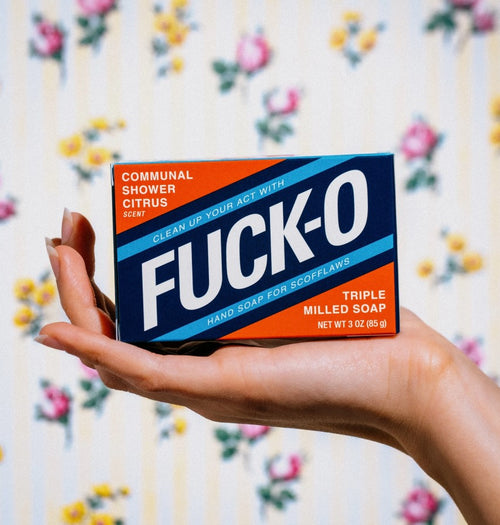A hand holding a rectangular bar of soap from Blue Dog Posters, featuring bold text that reads "FUCK O" against a background of floral-patterned wallpaper. The vintage-style package also mentions "COMMUNAL SHOWER CITRUS SCENT," "HAND SOAP FOR SCOFFLAWS," and "TRIPLE MILLED SOAP.