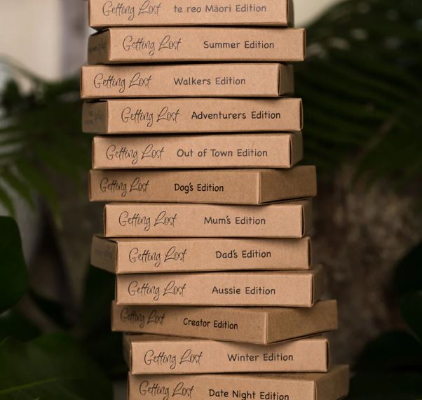 Several stacked Blue Dog Posters boxes showcase different editions from the "Getting Lost Cards" game series. Labels include "Summer Edition," "Walkers Edition," "Adventurers Edition," and more. Explore on foot with a different walk each time! The backdrop features lush green plants.