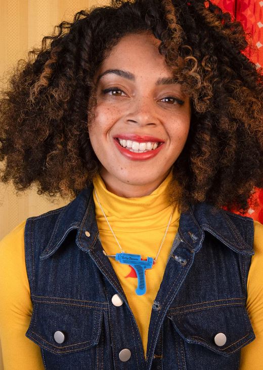 A person with curly hair smiles while wearing a yellow turtleneck and denim vest. A Blue Dog Posters Glue Gun Necklace, crafted from recycled acrylic, hangs from their neck. The backdrop features a yellow and red patterned wall.