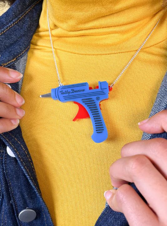 A person wearing a yellow top and denim jacket showcases a Glue Gun Necklace by Blue Dog Posters, crafted from recycled acrylic, featuring a blue and red laser gun pendant.