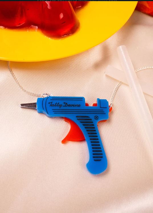 A Glue Gun Necklace by Blue Dog Posters, featuring a blue design and crafted from recycled acrylic, sits on a cream-colored surface. Nearby lie a translucent straw and red, jelly-like shapes.