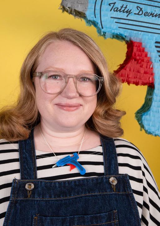 A person with long hair and glasses smiles at the camera, wearing a striped shirt and denim overalls. They're accessorized with a colorful Glue Gun Necklace crafted from recycled acrylic by Blue Dog Posters. The background is bright yellow, with part of a colorful piñata visible.