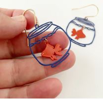 A close-up shows a hand holding Blue Dog Posters' Mini Goldfish Bowl Dangle Denz Earring, featuring a wire-frame fishbowl with an orange fish. Another similar earring is blurred in the background.