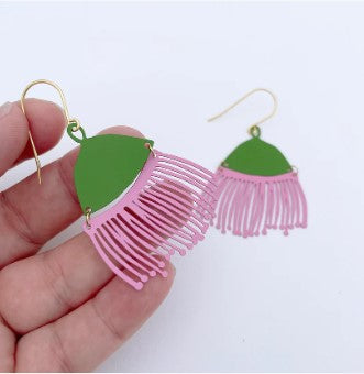A hand holds a pair of colorful, lightweight statement earrings named Denz Gum Blossom Pink & Lilac Earrings by Blue Dog Posters. The earrings are shaped like flowers with green tops and fringed pink petals hanging below. Each earring, made from recycled stainless steel, has a hook for wearing. The background is plain and white.