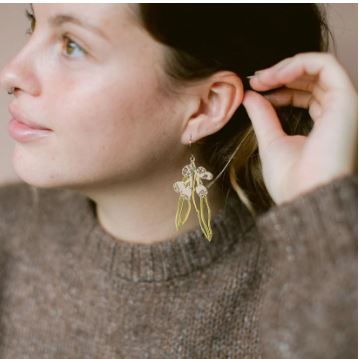 A person with light skin adjusts the Gumnut and Leaf Gold Silver Denz Earrings from Blue Dog Posters. They sport a brown knit sweater, have their hair pulled back, and stand against a softly blurred background.