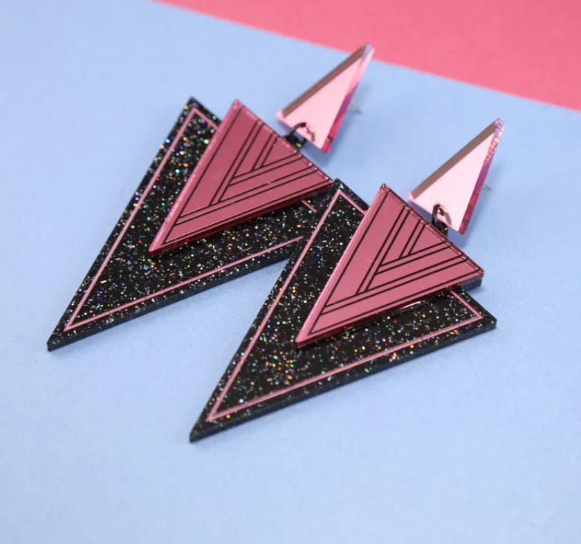 Introducing the Hannie Pink Black Dangle Earrings from Blue Dog Posters! These stunning handmade Art Deco earrings feature a striking geometric triangular design with a captivating blend of pink and black, accented with glitter. Displayed on a pastel pink and blue background, these unique earrings are available exclusively at Fox and Ginger.