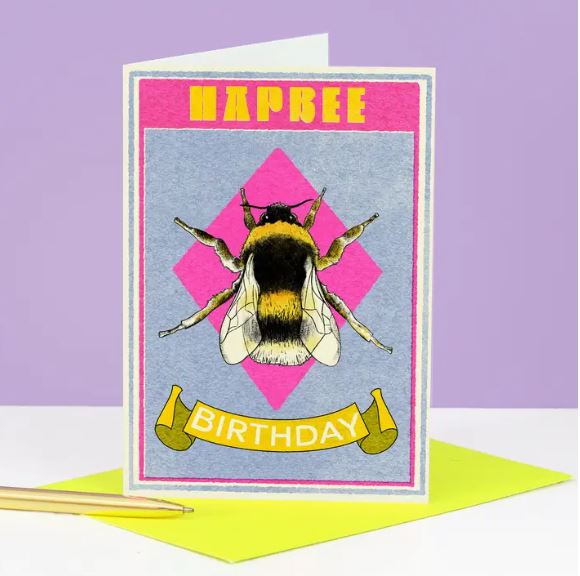 The Hapbee Birthday Card by Blue Dog Posters features a bee on a pink diamond background, with "Hapbee Birthday" at the top and bottom. A yellow envelope and pencil sit beside it against a purple backdrop.