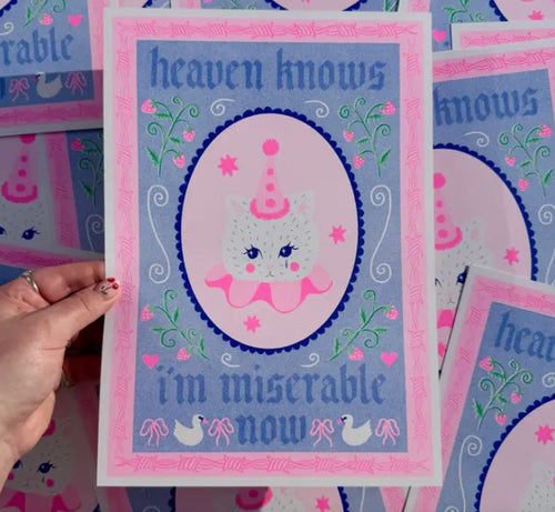 A hand holds the "heaven knows A4 Risograph" by Amy Hastings, featuring a sad cat in a party hat and ruff with decorative elements. The text says "heaven knows I'm miserable now," embellished with pastel heart and bird motifs. From Blue Dog Posters.